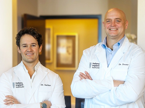 Dr. Surber and Dr. Drone, dental implant specialists at Indiana Implant Institute, smiling confidently in white lab coats.
